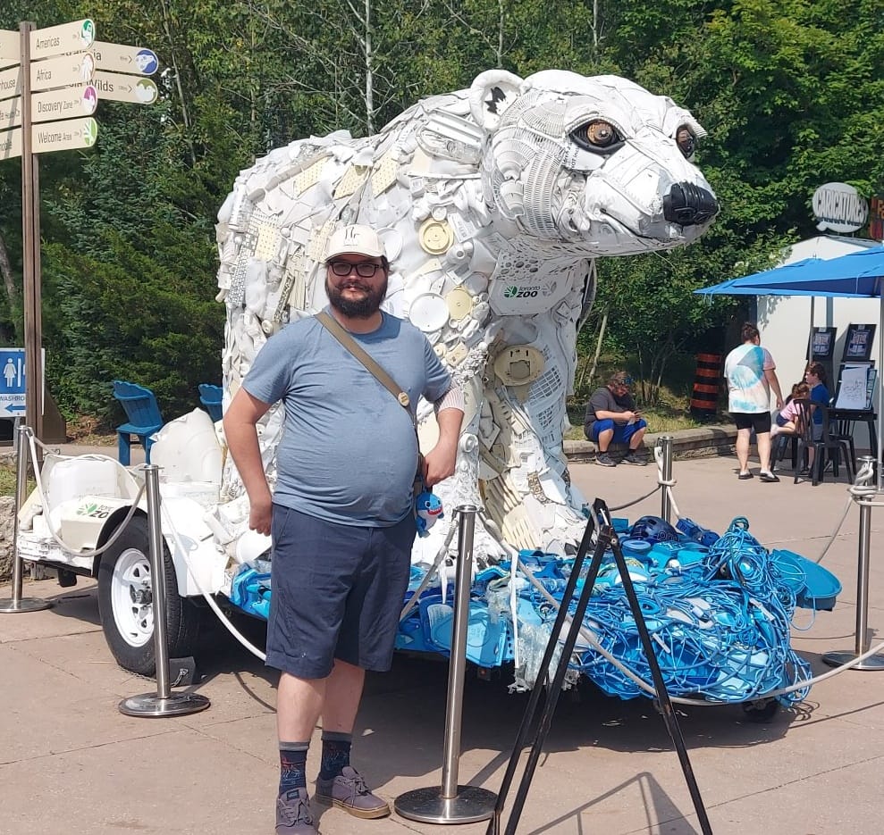 A man short hair and bushy facial hair stands in front of a large sculpture of a polar bear made out of plastic waste